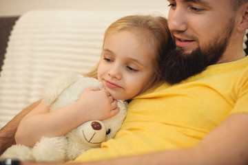 Beautiful little blonde girl leaning on her father's chest listeing what her father is reading. Bearded father embracing her little girl at home .