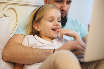 Close up of a lovely little blonde girl biting her finger smiling while her father is choding cartoons on his laptop in the bed . - obrazy, fototapety, plakaty