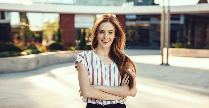 Side view portrait of a beautiful young female entrepreneur with red hair and freckles talking at phone outdoor near her workplace against a building.