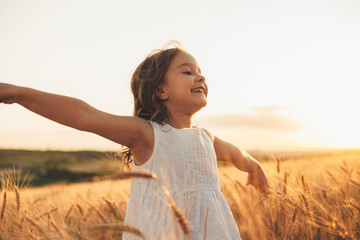 Beautiful little girl laughing and running with hand up in a wheat field against sunset. Freedom...