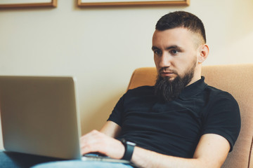 Young bearded man sitting in sofa and looking a laptop holding it on his legs at home. Male freelancer working at his laptop at home .