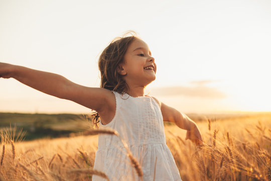 Beautiful little girl laughing and running with hand up in a wheat field against sunset. Freedom concept.