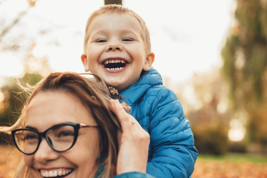 Lovely Little Kid Laughing While Sitting On His Mother's Back Outdoor Looking At Camera.