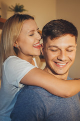 Close up portrait of a beautiful blonde woman embracing her boyfriend from and and touching his ear with the tongue laughing in the kitchen at home.