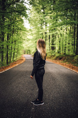 Young athletic sporty girl with long blonde hair standing on the asphalt road in the green forest.