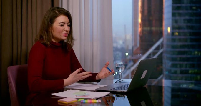 a brunette sits at a round table near a window. she talks on a video link through a laptop, puts stickers on paper, begins to write. behind the window you can see high-rise city buildings. close up