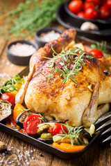 Baked whole chicken with vegetables and herbs close-up on a plate on a table.