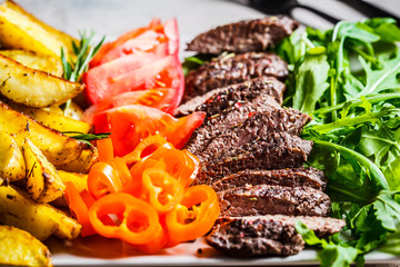 Chopped beef steak, baked potato wedges and vegetables on gray plate, gray background.