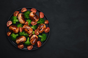 grilled sausages with tomatoes,  broccoli on black plate. dark background top view