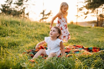 Little sisters: girls having fun in the summer forest, sitting on the blanket and smiling