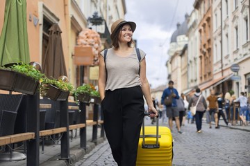 Traveling smiling mature woman tourist in hat with backpack and suitcase walking