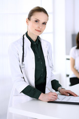 Doctor woman at work with patient and colleague at background. Physician filling up medical documents or prescription while standing in hospital reception desk. Data in medicine and health care