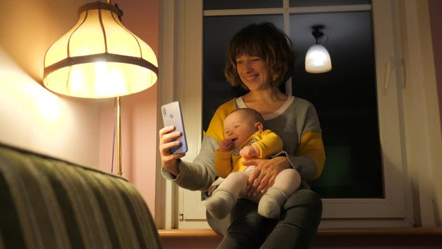 Mother with baby child enjoy take phone selfie pictures sitting on windowsill near lamp at home