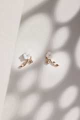 Subject shot of a pair of stud earrings isolated on the beige surface with gray shadows. Each of the earrings is made as a white flower, golden leaves and crystals.