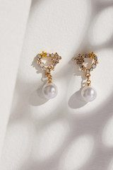 Subject shot of a pair of golden stud earrings on the beige surface with gray shadows. Each of the earrings is made as a heart-shaped flower crown with gems, pearls, crystals and a pearl pendant. 