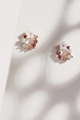 Subject shot of a pair of stud earrings isolated on the beige surface with gray shadows. Each of the earrings is made as a golden flower crown with gems, pearls and crystals. 