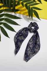 Subject shot of a blue satin hair scarf fixed on the scrunchie with white Paisley print. The scrunchie is isolated on the gray and yellow surface decorated with tropic leaves.