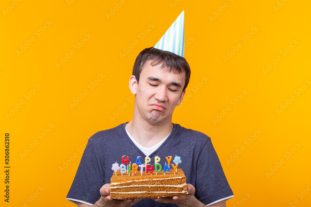 Wall mural Crazy cheerful young asian guy with glasses holding a burning candle in his hands and a congratulatory homemade cake on a yellow background. Birthday and anniversary celebration concept.