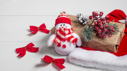 Toy snowman with Christmas present and red hat on a white wooden table.