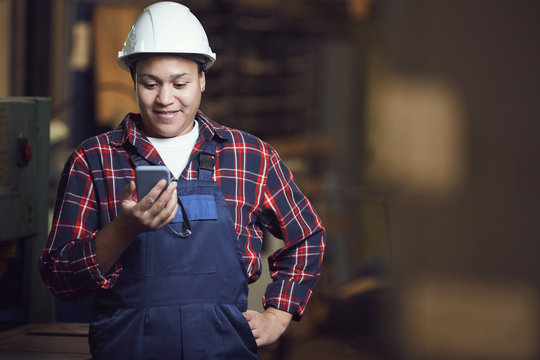 Waist up portrait of modern female worker looking at smartphone screen while standing by machine units in industrial plant, copy space