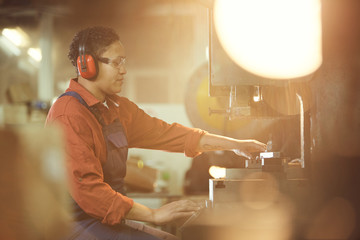 Side view portrait of modern African-American woman operating machine units while enjoying work at factory in sunlight, copy space