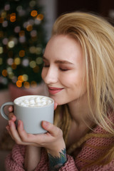 A large portrait of a beautiful attractive happy young sweet gently smiling blonde girl with dreadlocks in a pink cozy sweater with a Cup of coffee with marshmallows on a background of bokeh.