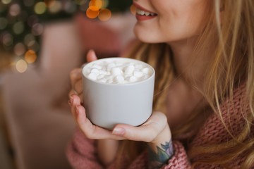 A large portrait of a beautiful attractive happy young sweet gently smiling blonde girl with dreadlocks in a pink cozy sweater with a Cup of coffee with marshmallows on a background of bokeh.