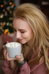 A large portrait of a beautiful attractive happy young sweet gently smiling blonde girl with dreadlocks in a pink cozy sweater with a Cup of coffee with marshmallows on a background of bokeh.