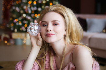 A large portrait of an attractive happy cute smiling blonde girl with dreadlocks in a pink cozy sweater with a Christmas tree toy in her hand on the background of bokeh, garlands and a sofa.