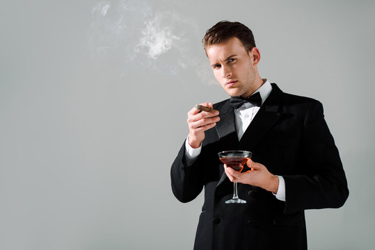 Rich Man In Suit With Bow Tie Holding Glass With Alcohol Drink And Cigar Isolated On Grey