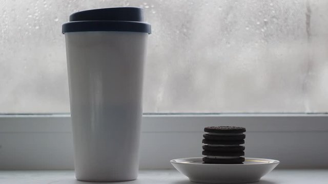 A Cup with a lid with coffee stands on the windowsill, next to a plate are cookies stacked in a pile. A woman's hand appears from outside the frame and takes the cookie and then returns it bitten off