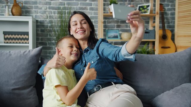 Loving mom taking selfie with adorable son using smartphone camera hugging child showing thumbs-up and v-sign hand gestures smiling feeling love. People and gadgets concept.