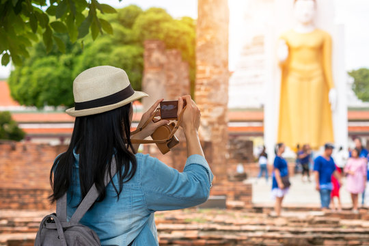 The back of an Asian woman wearing a hat and denim shirt taking pictures of Buddha statues