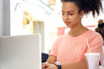 In the Cafe. Young girl in glasses sitting at table with milkshake working on laptop concentrated close-up