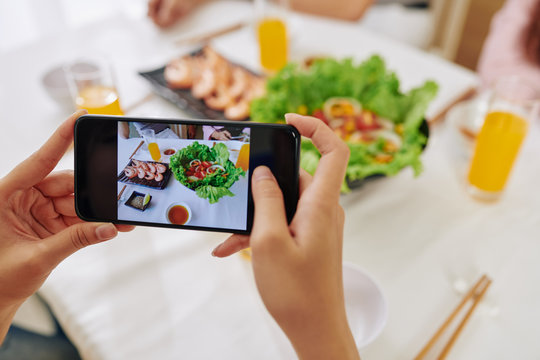Close-up image of woman taking photos of shrimps and vegetable salad on dinner table for her blog