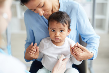 Young mother holding her baby boy while doctor listening to his heartbeat with stethoscope at...