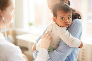 Young mother holding her little child on her hands while female doctor examining him with stethoscope at hospital