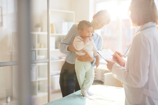 Female Doctor In White Coat Standing And Writing A Treatment While Mother Holding Her Baby During Their Visit At Hospital