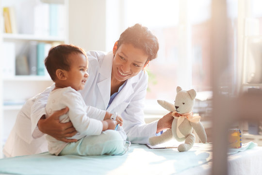 Young female doctor in white coat showing toy to her little patient and playing with him at hospital