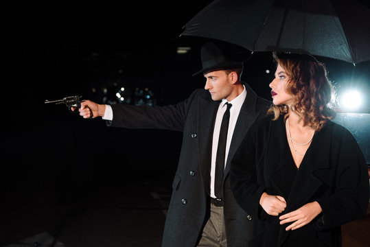 Side View Of Dangerous Man In Hat Holding Gun Near Attractive Girl And Retro Car On Black