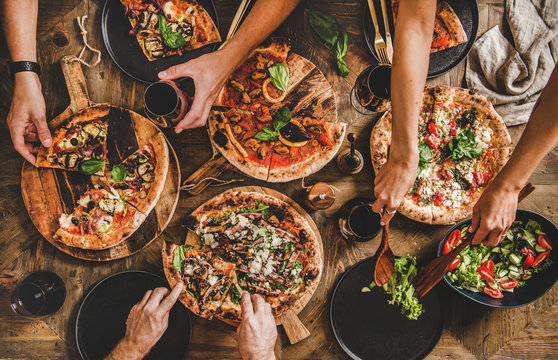 Family or friends having pizza party dinner. Flat-lay of people cutting and eating Italian pizza and drinking red wine from glasses over wooden table, top view. Fast food lunch, gathering, celebration