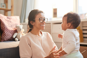 Happy young mother in eyeglasses talking to her little child in the room at home