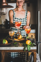 Young smiling woman in checkered dress holding Aperol Spritz aperitif drink with orange in hands,...