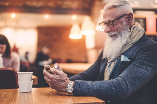 Happy trendy man drinking coffee and using smartphone in bar cafeteria indoor - Fashion senior guy having fun with web app - Technology trends and joyful elderly lifestyle concept - Focus on his hand