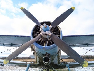 A four bladed black propeller on the nose of a green biplane aircraft against a blue sky