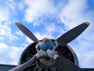 A four bladed black propeller on the nose of a green biplane aircraft against a blue sky