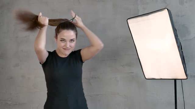 Caucasian Girl in a black t shirt long straight hair brown haired against the background of a gray wall and a lamp throws her head up collects her hair in ponytail fooling around in front of camera