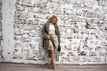 Fashion woman posing at the background street brick wall, dressed modern clothes. Female model wearing faux fur coat, tutleneck, skirt and bag