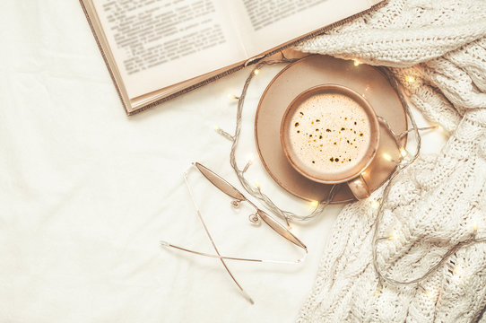 Coffee Cup With Foam, Reading Glasses, An Open Book And A White Sweater With A New Year's Garland And Lights On A White Bedsheet.  Top View.  Flat Lay