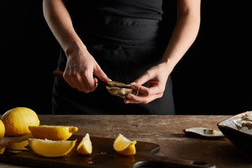cropped view of woman holding knife while opening oyster near lemons on cutting board isolated on black
